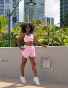 Woman in pink athletic Love fitness eloise sports bra and sweet heart sweatshorts in the colorway strawberry cream posing outdoors against a cement wall with palm trees and tall building in background.