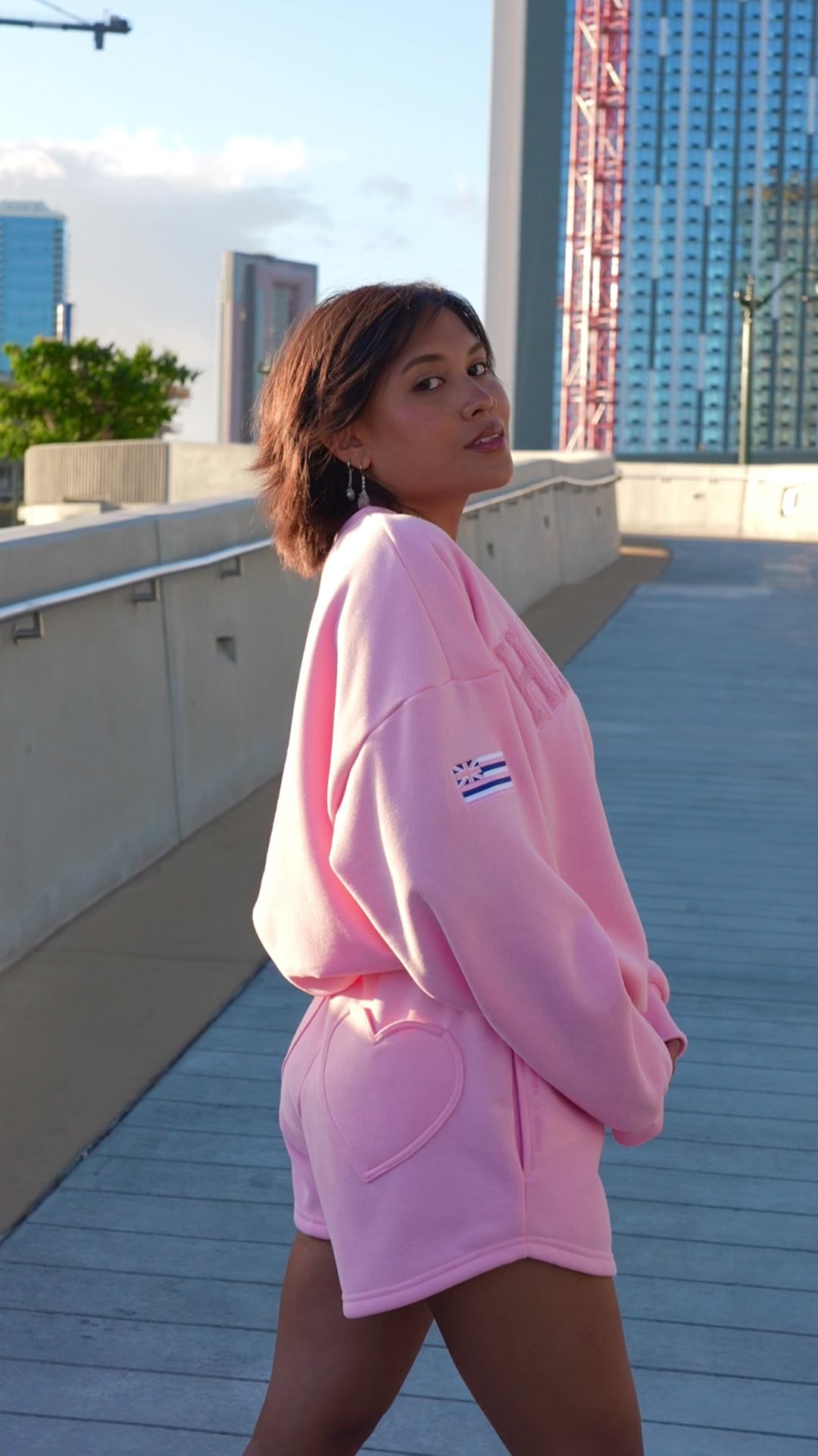 Person wearing Love Fitness State of aloha sweatshirt pink 'Hawaii' sweatshirt and shorts set, standing outdoors. Colorway is called strawberry pink matching with love fitness sweet heart sweatshorts in the same color pink.