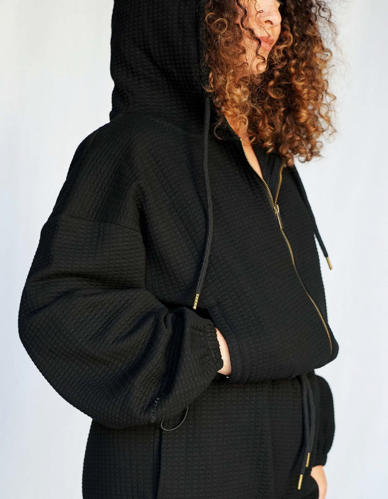 Love Fitness Phaedra Jacket in the color black. Waffle texture with a zipper, oversized hood and drawstrings on the side to adjust