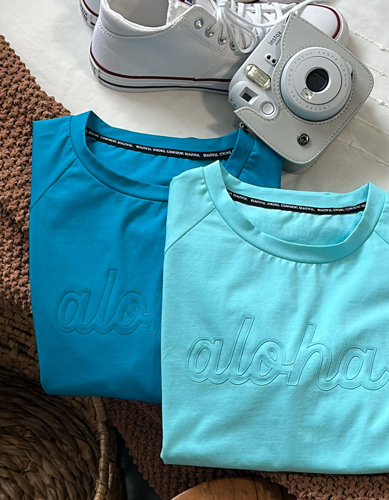 Love Fitness Aloha Tee in the color sweet tart and shave ice. Flatlay on the bed.