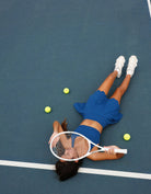 Person lying on a tennis court with a racket and balls. Person wearing Love Fitness Bodhi Active Skirt for tennis outfit on a tennis court. In the colorway blue opal.