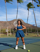 Woman in blue tennis outfit holding a racket on a tennis court with palm trees and mountains in the background. Person wearing Love Fitness Bodhi Active Skirt for tennis outfit on a tennis court. In the colorway blue opal.
