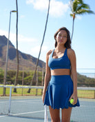 Woman wearing Love Fitness Eloise Sports bra in the colorway blue opal holding a tennis racket on a tennis court with mountains in the background