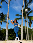 Woman in blue athletic wear standing in a tropical setting with palm trees and a clear sky. Wearing love fitness effortless leggings in the colorway blue opal