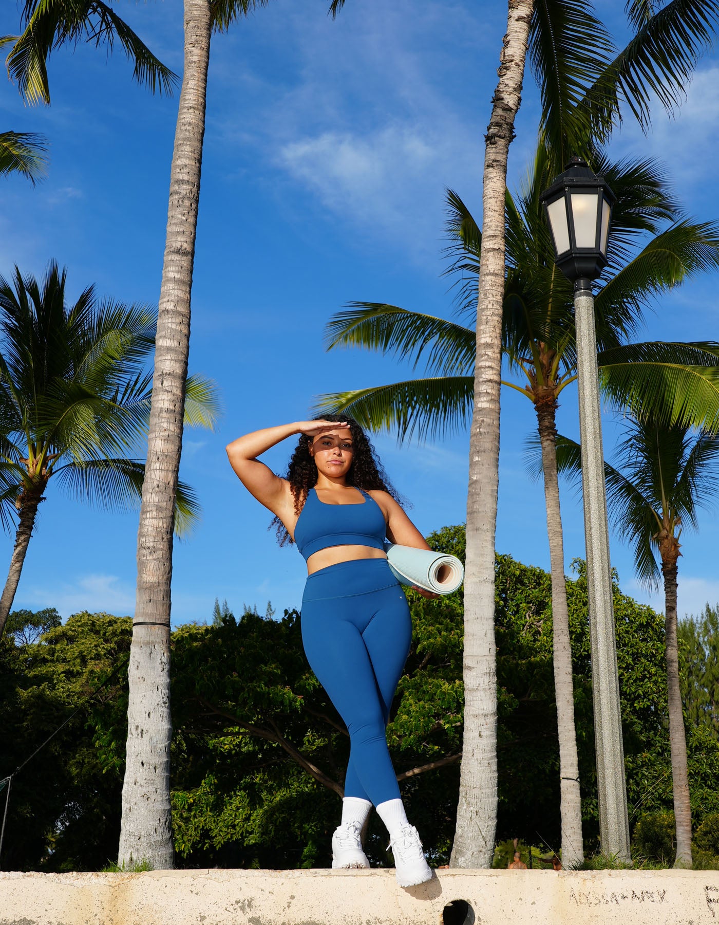 Woman in blue athletic wear standing in a tropical setting with palm trees and a clear sky. Wearing love fitness effortless leggings in the colorway blue opal
