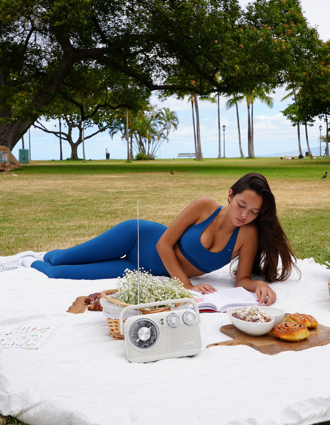 Woman wearing love fitness effortless leggings in the colorway blue opal swimsuit lying on a picnic blanket with a radio and food items in a park.