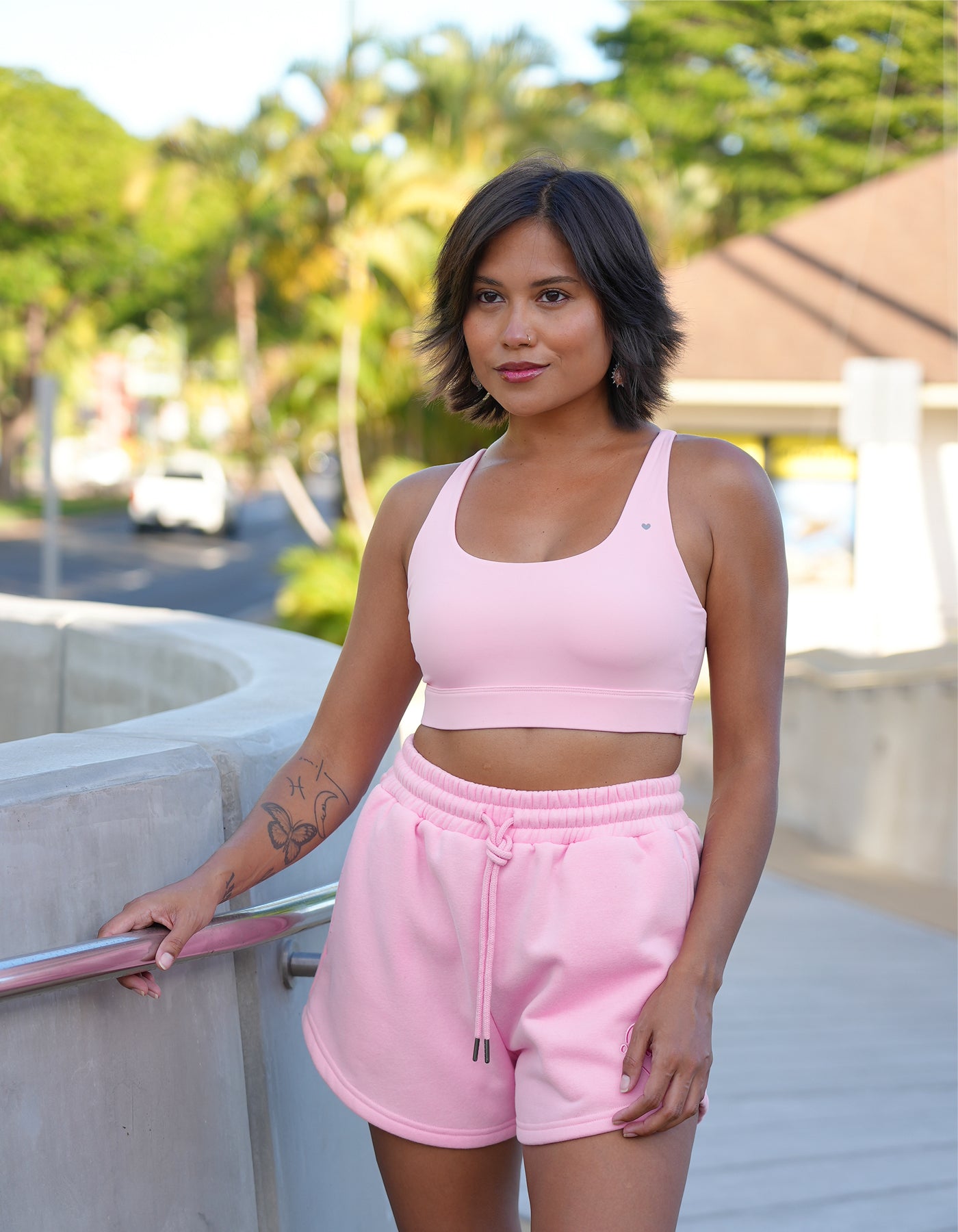 Love Fitness Eloise Sports bra in Strawberry Cream Pink. Model is standing on paved path holding side rail wearing a pink sports bra and pink sweatshorts with pockets and drawstring.