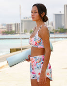 Woman is wearing  Love Fitness Floral Fable Runners holding a yoga mat by a waterfront with buildings in the background.