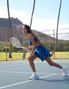 Woman playing tennis on a court with mountains in the background. Person wearing Love Fitness Bodhi Active Skirt for tennis outfit on a tennis court. In the colorway blue opal.