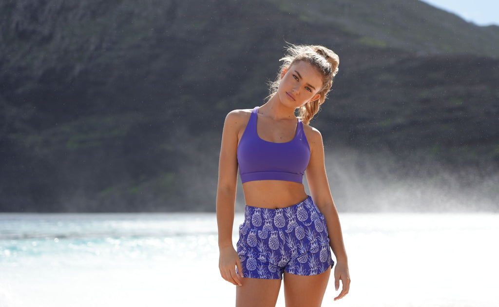 Pink & Purple Pineapple Collection with woman on beach wearing the new Pineapple runners and leilani sports bra in the color lavender