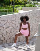 Woman in pink athletic Love fitness eloise sports bra and sweet heart sweatshorts in the colorway strawberry cream posing outdoors against a stone wall.
