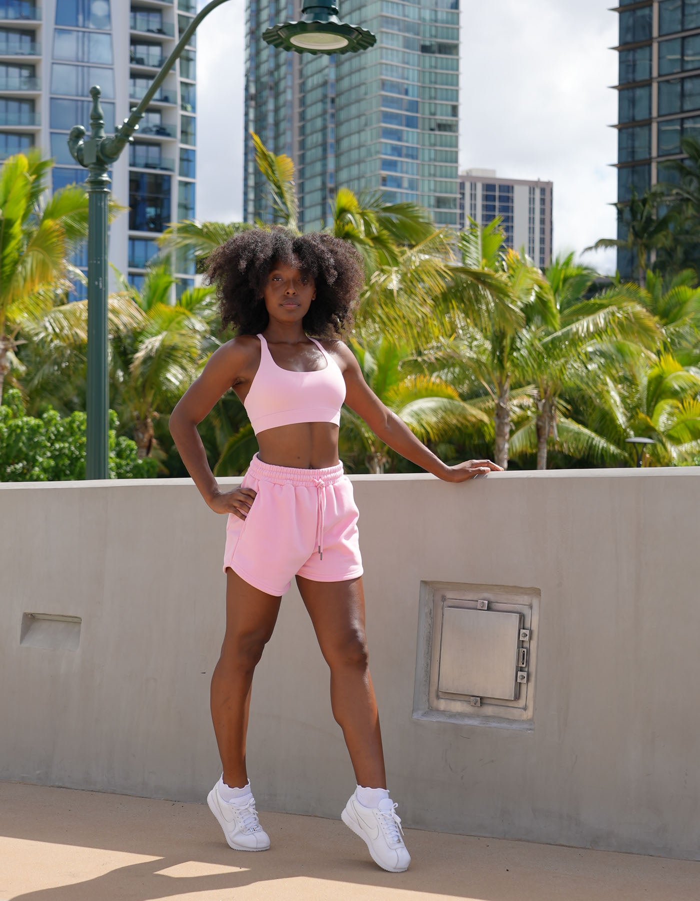 Woman in pink athletic Love fitness eloise sports bra and sweet heart sweatshorts in the colorway strawberry cream posing outdoors against a cement wall with palm trees and tall building in background.
