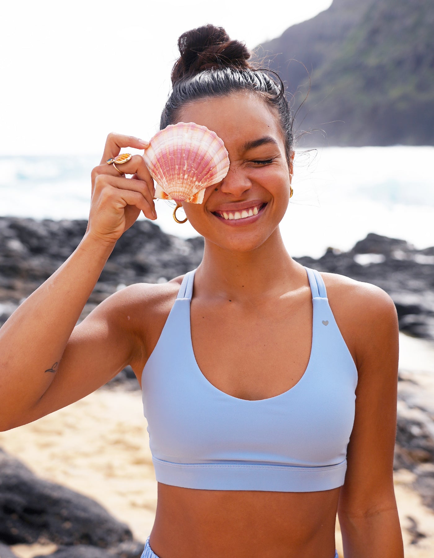 Love Fitness Johana Sports Bra in the color tidal mist. Woman holding a pink shell to her eye on a beach