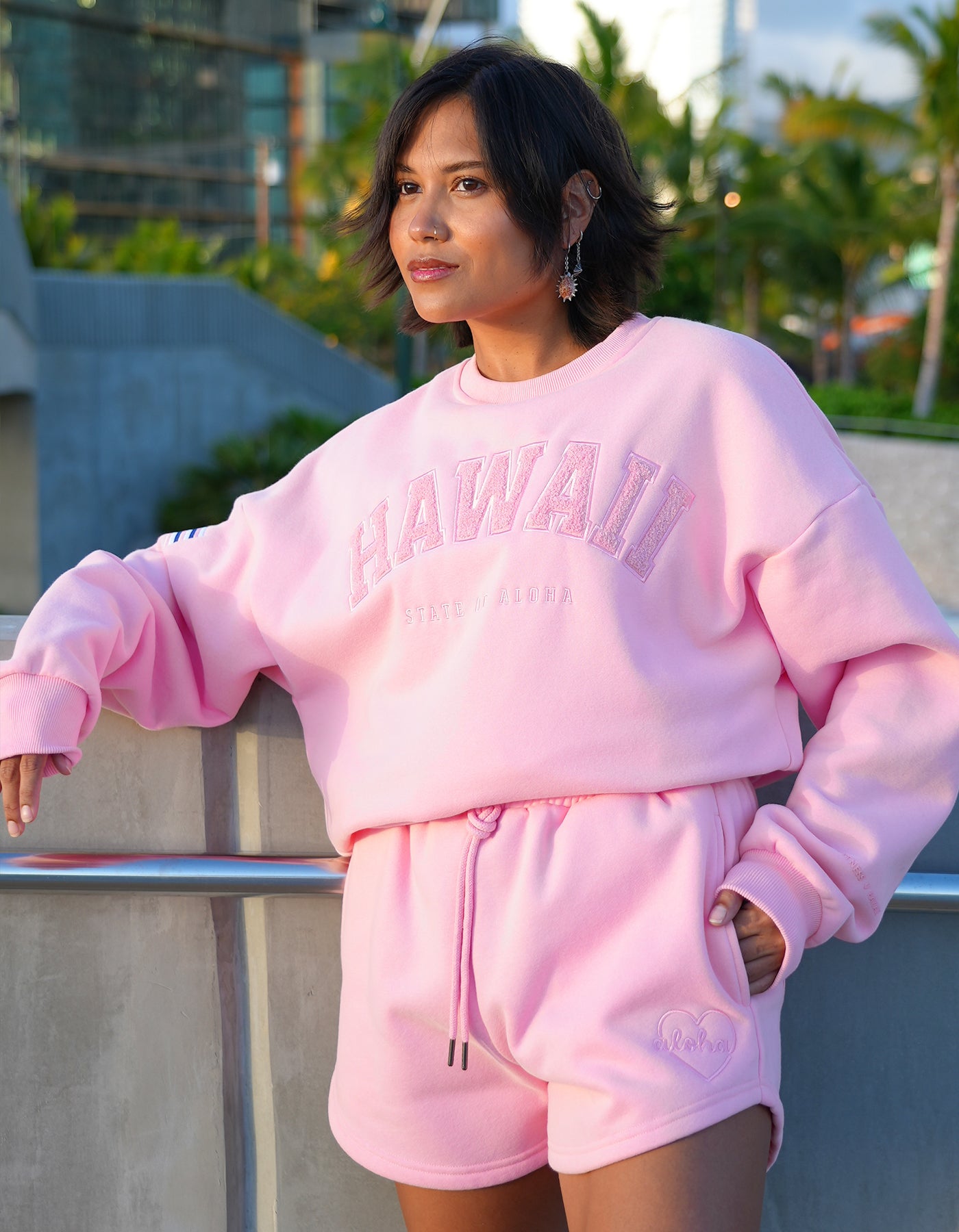 Person wearing Love Fitness State of aloha sweatshirt pink 'Hawaii' sweatshirt and shorts set, standing outdoors. Colorway is called strawberry pink matching with love fitness sweet heart sweatshorts in the same color pink.