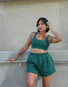 Woman wearing headphones wearing love fitness eloise sports bra and sweet heart sweat shorts in the color evergreen standing with one foot against the wall learning back with hand spread out holding the railing on the wall.