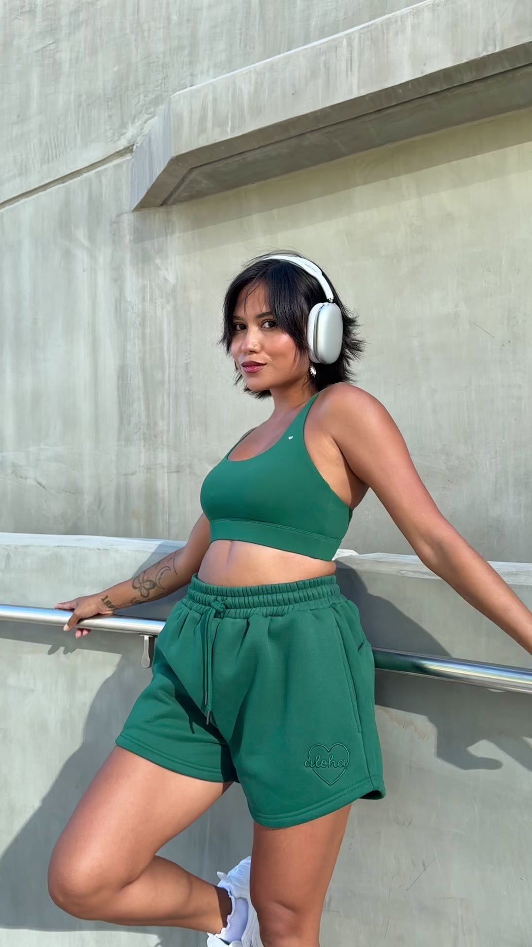 Woman wearing headphones wearing love fitness eloise sports bra and sweet heart sweat shorts in the color evergreen standing with one foot against the wall learning back with hand spread out holding the railing on the wall.