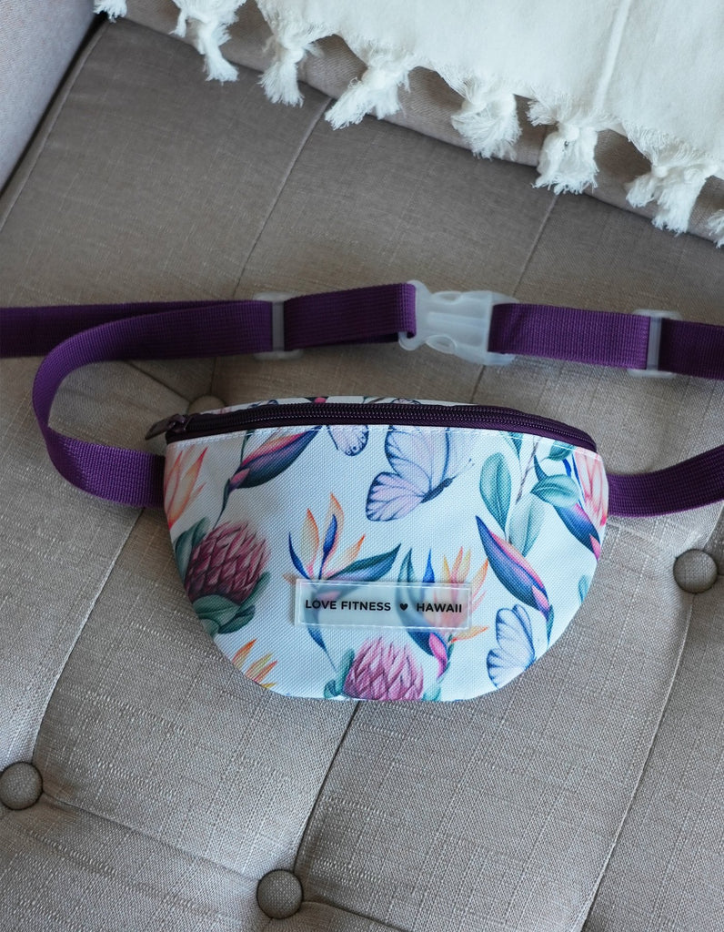 Love Fitness Island Dreams Fanny Pack with an all over print with island flowers and butterflies. Straps in a dark purple and compartment that fits all the on the go essentials