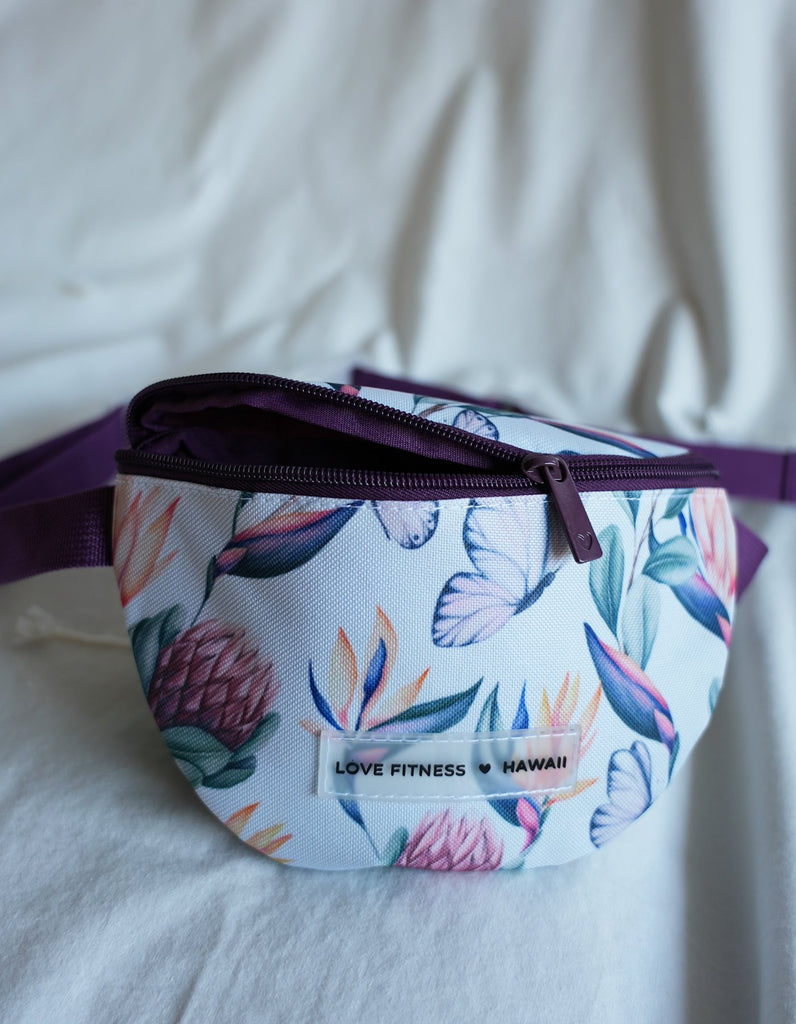 Love Fitness Island Dreams Fanny Pack with an all over print with island flowers and butterflies. Straps in a dark purple and compartment that fits all the on the go essentials. Dark purple zipper and zipper pull that is moder nand elegagnt